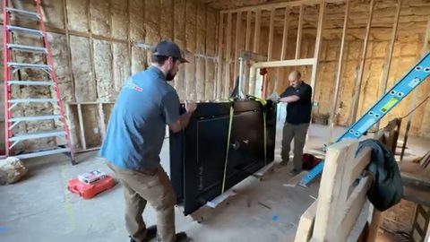 crew maneuvering a large black gun safe with straps and skates in a framed house