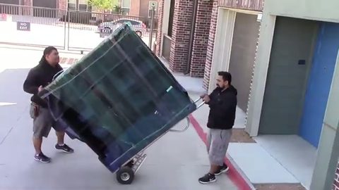 two movers wheeling a shrink-wrapped bookcase on a hand truck outside apartments, face visible
