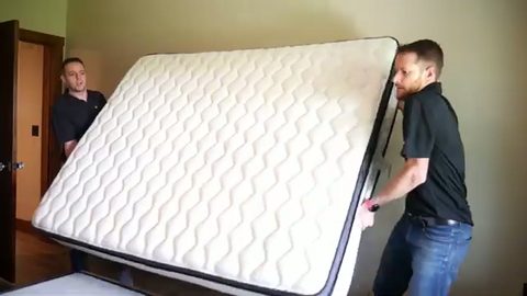 two movers lifting a mattress onto a bed in a bedroom, face visible